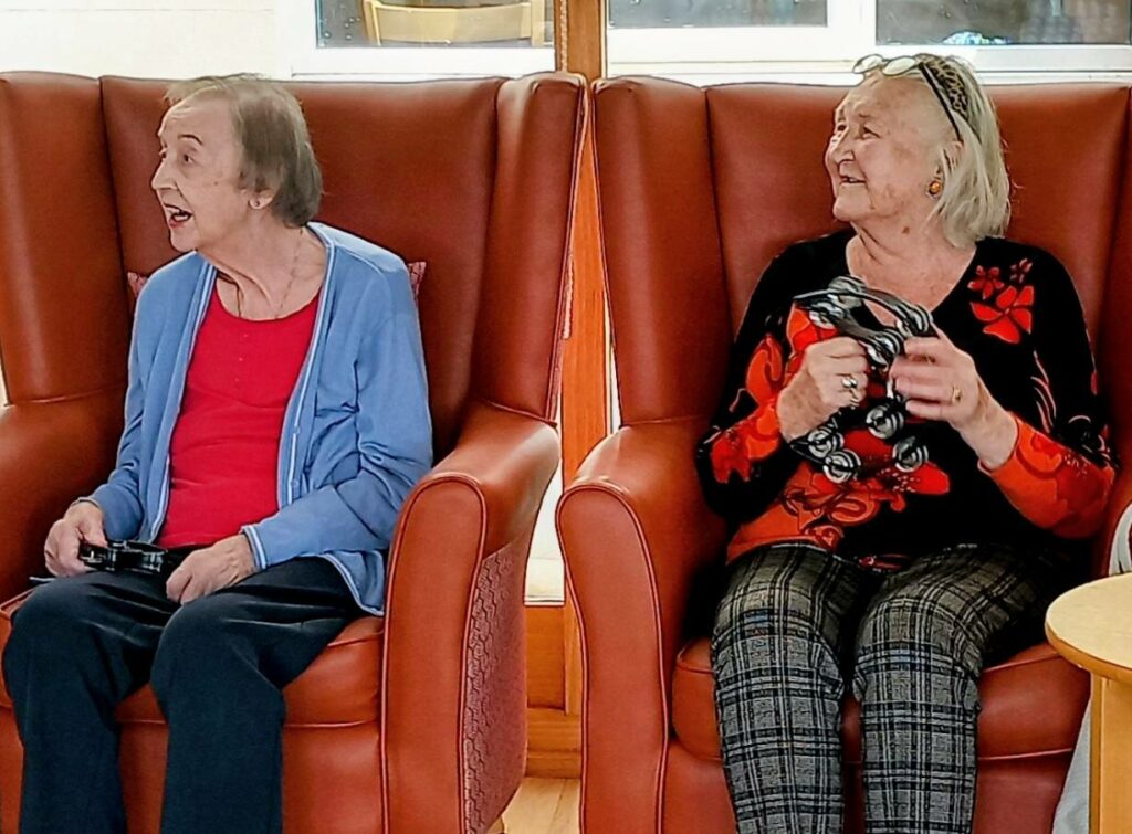 Two elderly women sit side by side in red armchairs, each holding a video game controller; one looks straight ahead while the other smiles at her companion.