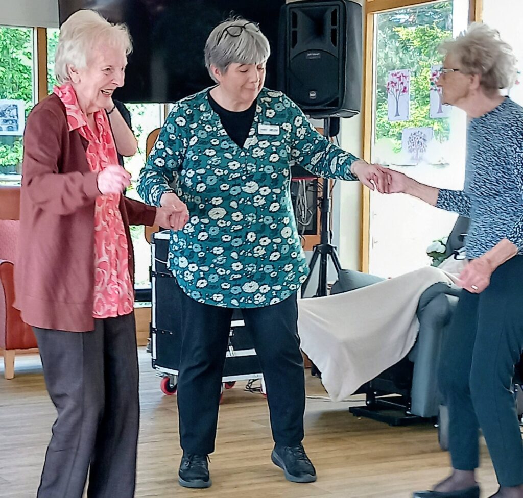 Three older women stand indoors holding hands, seemingly dancing or taking part in an activity together in a bright room adorned with artwork on the walls.