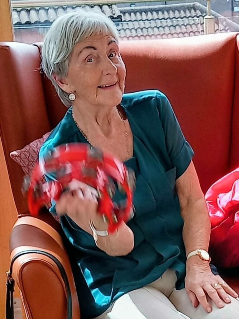An older woman with short grey hair sits in an armchair, smiling and holding a red tambourine in her right hand.