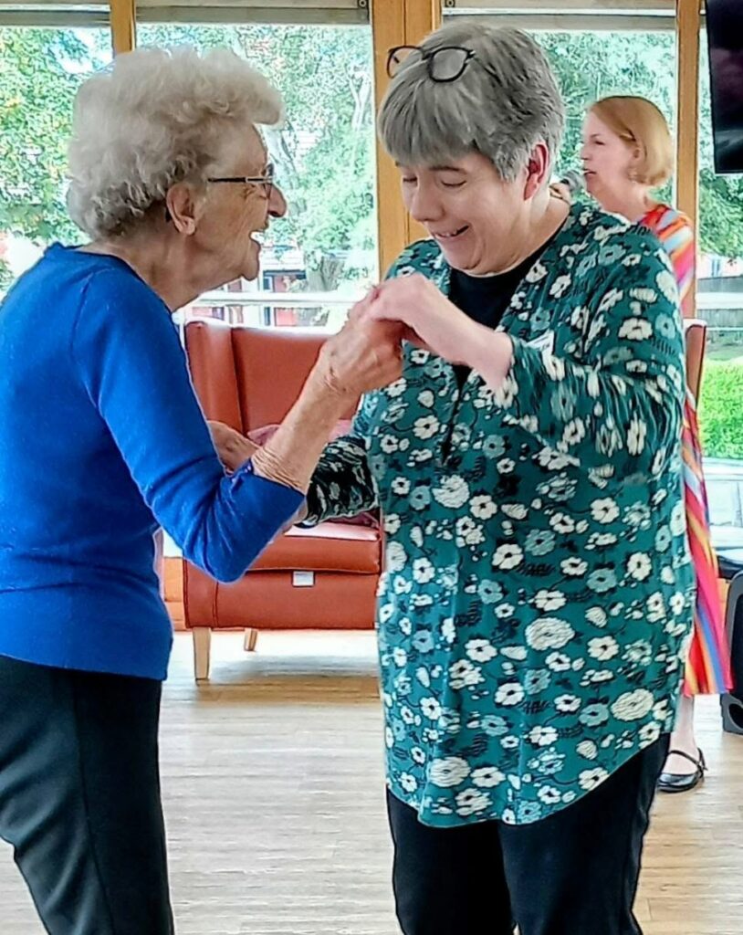 Two older women smile and hold hands while dancing indoors at Lear House, celebrating a special occasion, with another woman in colourful clothes in the background.