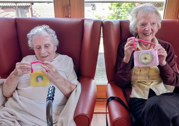 Two elderly women seated in armchairs chatting and holding up colourful, handmade knitted purses smile indoors by a window.