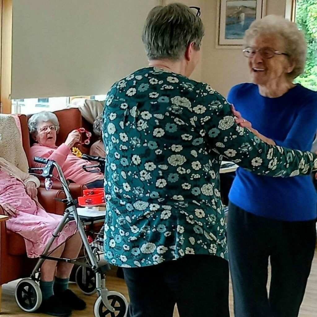 Two elderly women dance together whilst another woman sits on a sofa nearby with a zimmer frame beside her in a cosy, well-lit room.