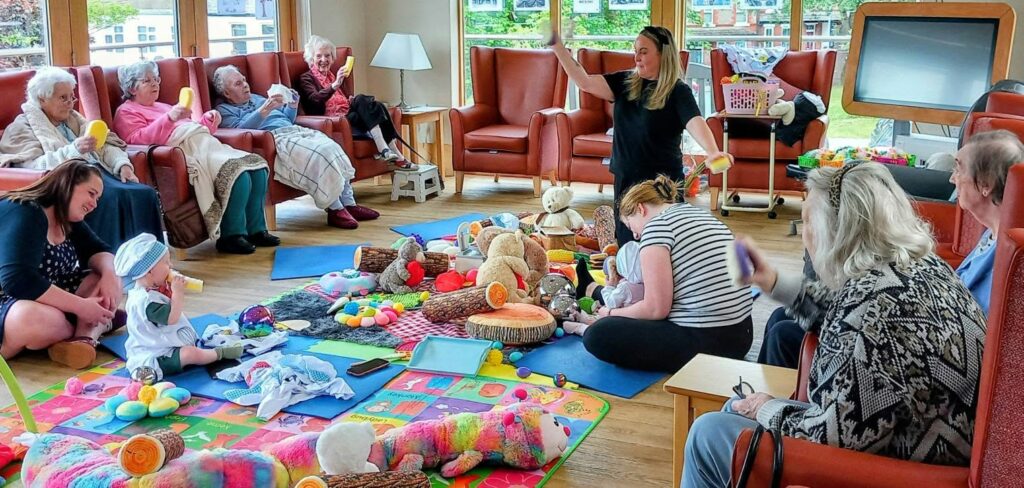 A group of elderly people sit in a circle watching babies and toddlers enjoying sensory play on mats with toys in a brightly lit community room. An adult, leading Boogie Baby, stands and gestures in the centre.