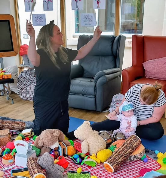 A woman lifts two sticks while another woman sits on the floor holding a baby. Toys, soft toys, and play food for sensory activities are scattered across a mat in the foreground.