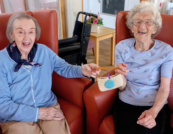 Two elderly women sit in armchairs, smiling and having a natter as they hold a small handmade bag together. A wheelchair and a table with a vase of flowers are in the background.
