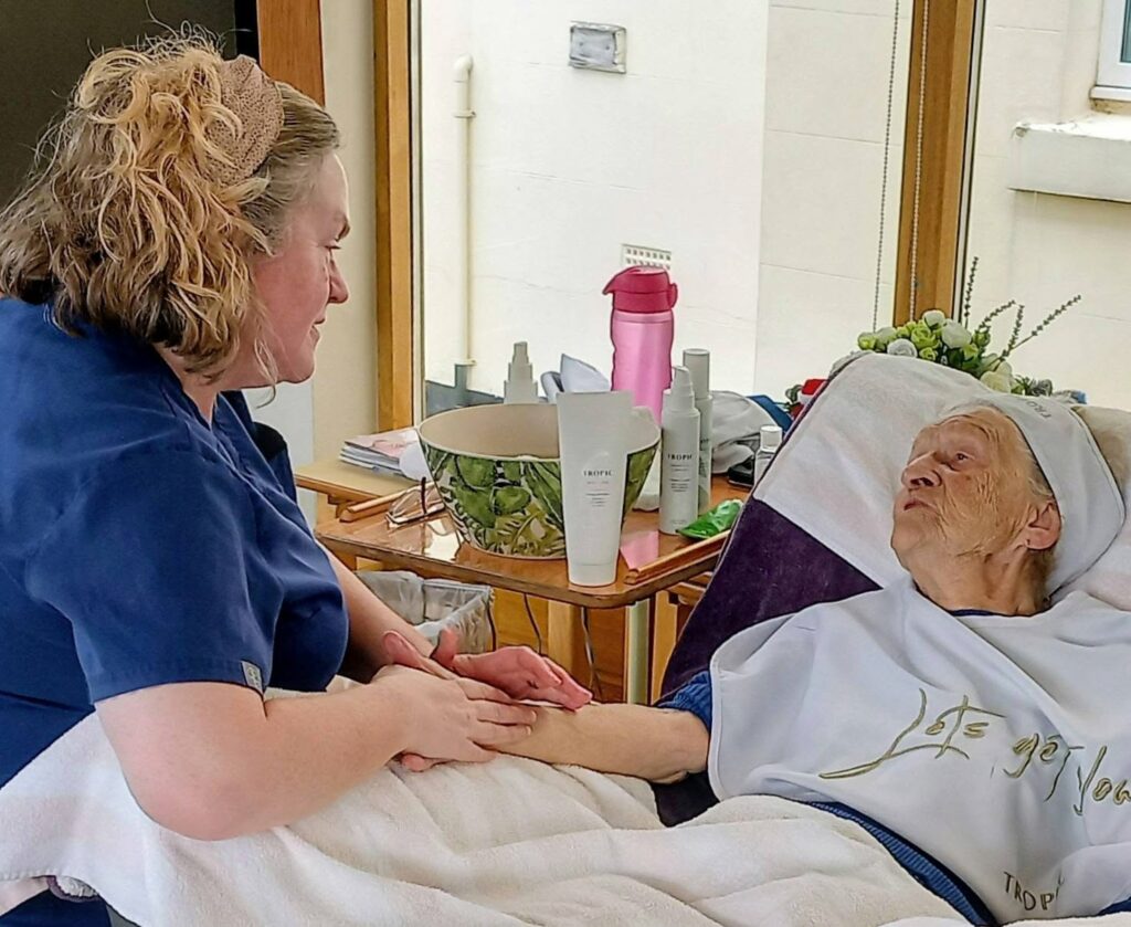 A carer in blue scrubs holds and massages the hand of an elderly woman lying in bed. Skincare products and a water bottle are on the nearby table.