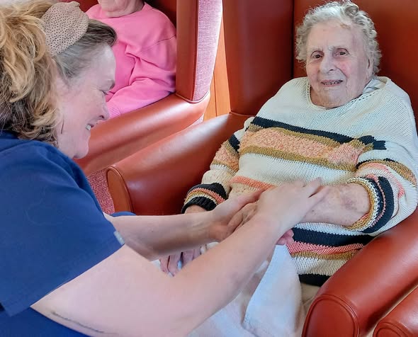A carer smiles as she holds the hands of an elderly woman seated in a chair, with another person sitting in the background.