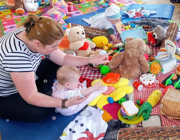 An adult and a baby sit on a mat surrounded by colourful toys, cuddly animals, and play food items, engaging in sensory play during a lively Boogie Baby activity.
