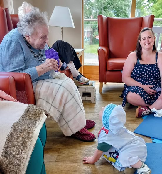 An elderly woman sits in an armchair holding a purple object, while a baby enjoys sensory play on the floor and a smiling woman in a spotty dress nearby creates an intergenerational musical moment.