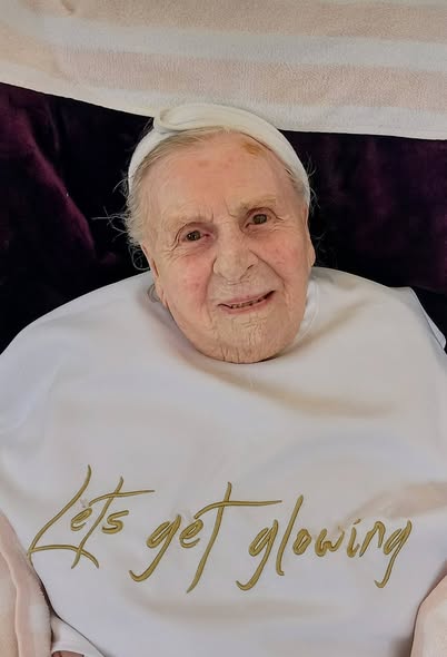Elderly woman wearing a headband and a white bib with 'Let's get glowing' written on it, lying on a dark surface.
