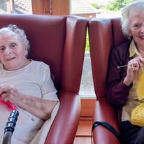 Two elderly women sit in armchairs by a window, chatting and knitting with smiles on their faces. A walking stick rests beside one of them.