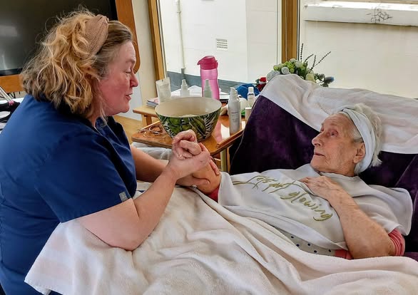 A carer holds hands with an elderly woman lying in bed, surrounded by personal care items and covered with a blanket.