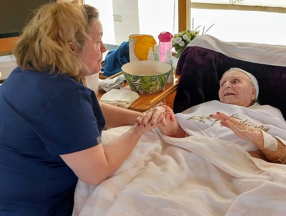 A nurse holds the hands of an elderly woman lying in a hospital bed, both making eye contact in a well-lit room with medical and personal items close by.