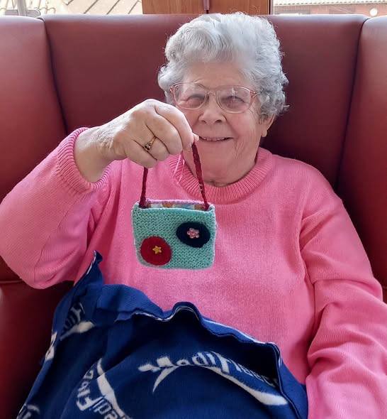 An elderly woman in a pink jumper smiles as she holds up a small, handmade knitted bag adorned with two floral patterns—proof that knitting can quietly change the world.