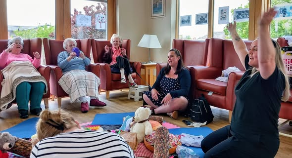 A group of elderly women and two younger adults sit in a circle indoors, some with hands raised, taking part in intergenerational music and sensory play, surrounded by blankets and toys.