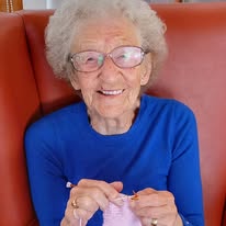 Elderly woman wearing glasses and a blue shirt sits in a red armchair, smiling as she knits with light-coloured wool and chats about making a difference in the world.