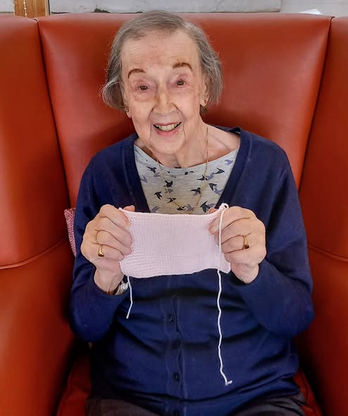 An elderly woman sits in a red armchair, smiling and chatting as she holds up a piece of pale pink knitting.