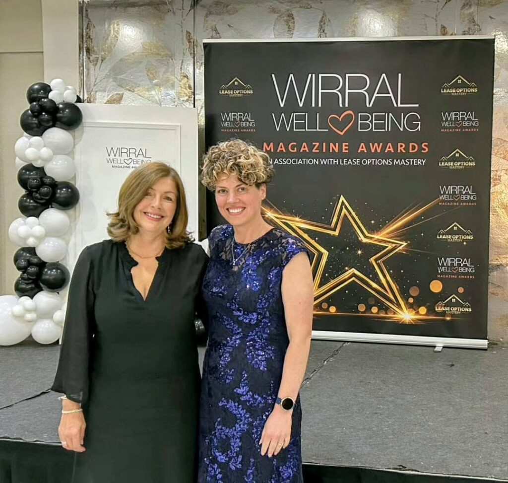 Two women pose and smile in front of a Wirral Well-Being Magazine Awards banner at an indoor event decorated with black and white balloons.