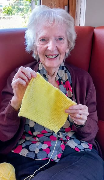 An elderly woman sits on a red bench, smiling and holding up a yellow knitted square—proof that knitting could be her way of changing the world, one stitch at a time.