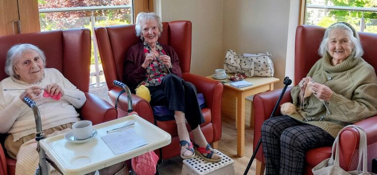 Three elderly women sit in armchairs, smiling and knitting together, chatting happily in a bright room with large windows, a table with cups, and a bag nearby.