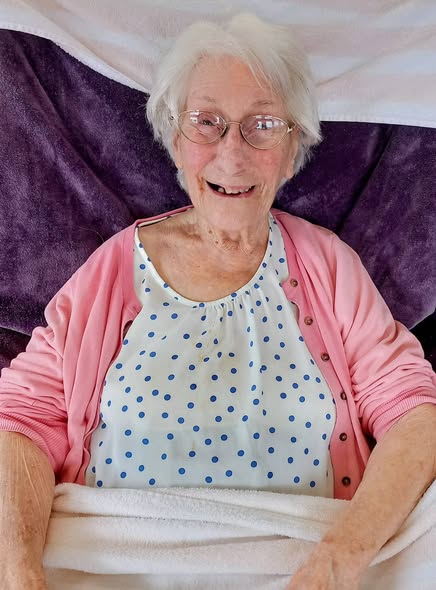 An elderly woman with white hair, spectacles, and a pink cardigan smiles whilst reclining on a bed covered with white and purple blankets.