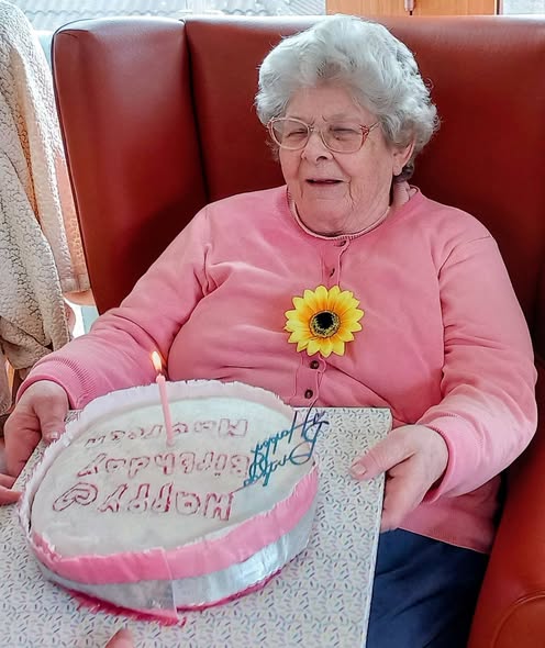 Elderly woman in spectacles and a pink cardigan holds a birthday cake with a single lit candle and 'Happy Birthday Maureen' written on top, marking a special celebration at Lear House.