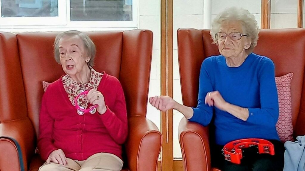 At Lear House, two elderly women sit in red armchairs; one, dressed in red, is singing whilst the other, in blue, plays a tambourine during a special celebration—Happy Birthday Maureen!.