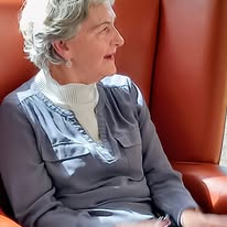 Maureen, seated in an orange chair at Lear House, wears a grey blouse over a white polo neck, looking to her left during the birthday celebration.