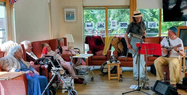 Two musicians play a washboard and banjo for a group of elderly residents seated in a care home lounge.