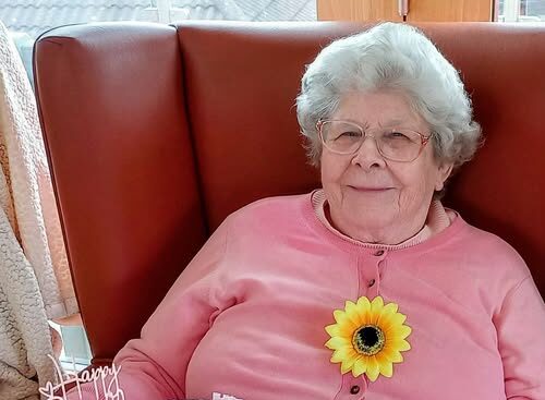 An elderly woman in a pink jumper, wearing spectacles and a sunflower brooch, holds a decorated birthday cake reading Happy Birthday Maureen at the special celebration in Lear House.