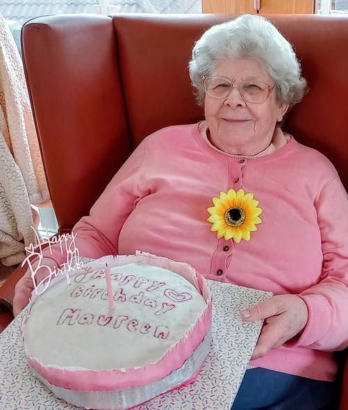 An elderly woman in a pink jumper, wearing spectacles and a sunflower brooch, holds a decorated birthday cake reading Happy Birthday Maureen at the special celebration in Lear House.