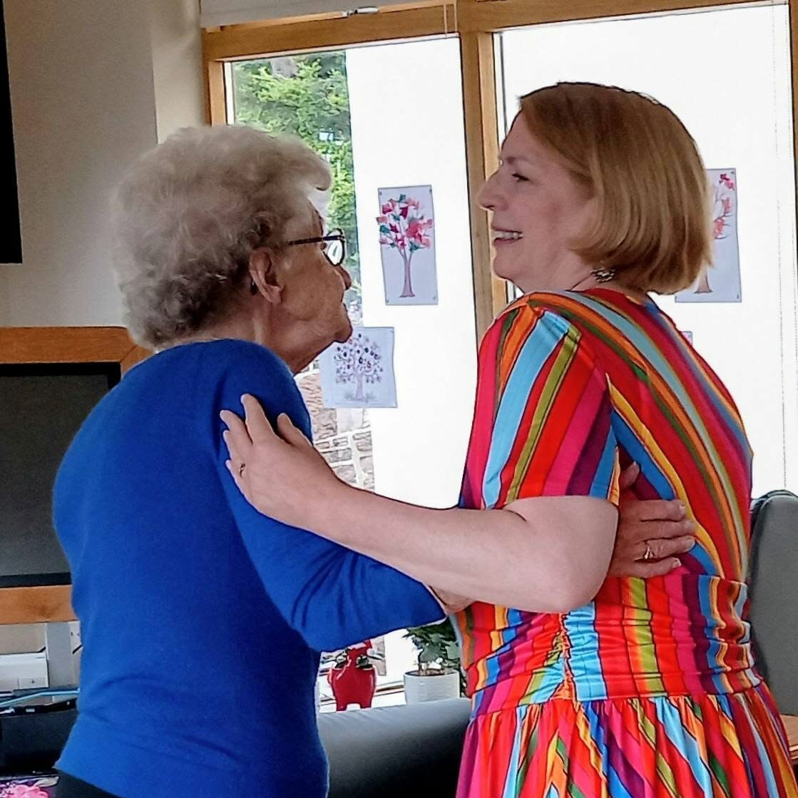 Two women stand facing one another indoors at Lear House, smiling and holding each other’s arms. One is dressed in a blue top, the other in a colourful striped dress—celebrating a special occasion. Happy Birthday Maureen!.