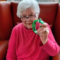 An older woman with white hair and glasses, dressed in a pink top, sits in a red chair at Lear House, holding up a green item towards the camera for Maureen’s special celebration.