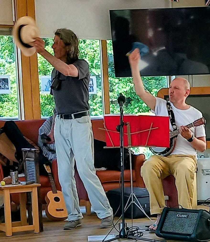 Two men perform indoors; one stands with a hat raised, while the other sits playing a banjo. Musical instruments and equipment can be seen around them.