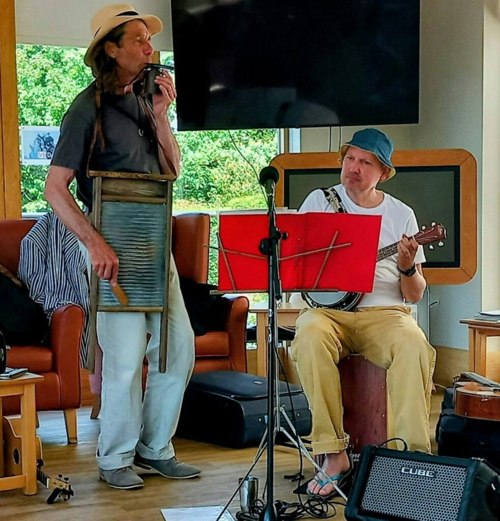 Two men perform indoors; one plays a washboard and harmonica, while the other plays a banjo. Music stands, microphones, and amplifiers are arranged around them.