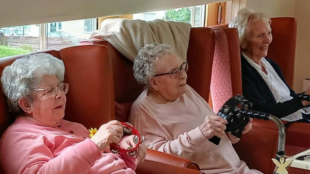 At Lear House, three elderly ladies relax in armchairs; two are holding game controllers, while one is knitting. A walking frame sits nearby. It’s a special celebration—Happy Birthday, Maureen!.
