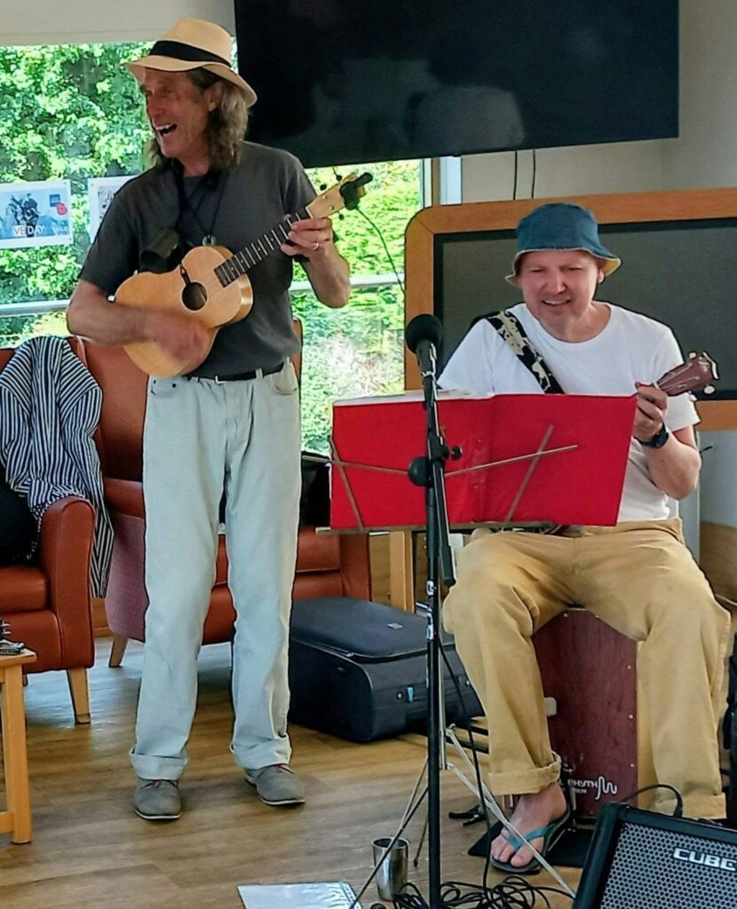 Two men perform indoors; one stands playing a ukulele and singing, while the other sits playing both a cajón and a ukulele. Both are wearing hats.