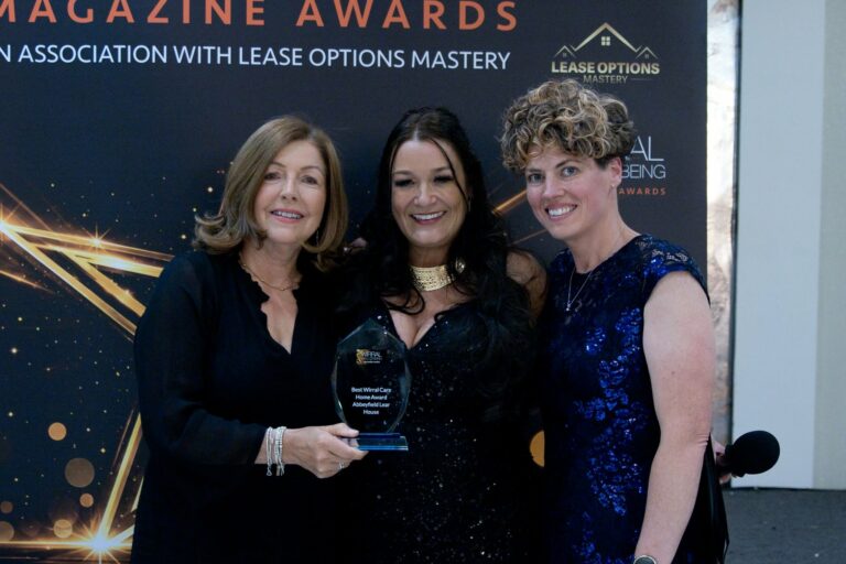 Three women in formal attire stand together, smiling, with one holding an award at an event featuring a black and gold banner in the background.