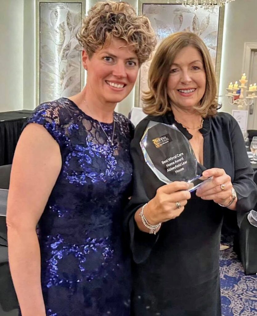 Two women stand together indoors, one holding a glass award. Both are smiling and dressed in formal attire.