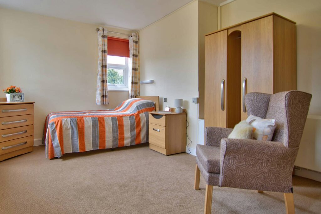 A neatly made single bed with an orange-striped cover, wooden furniture, an armchair, and a window with patterned curtains create a cosy home in this tidy, carpeted room.