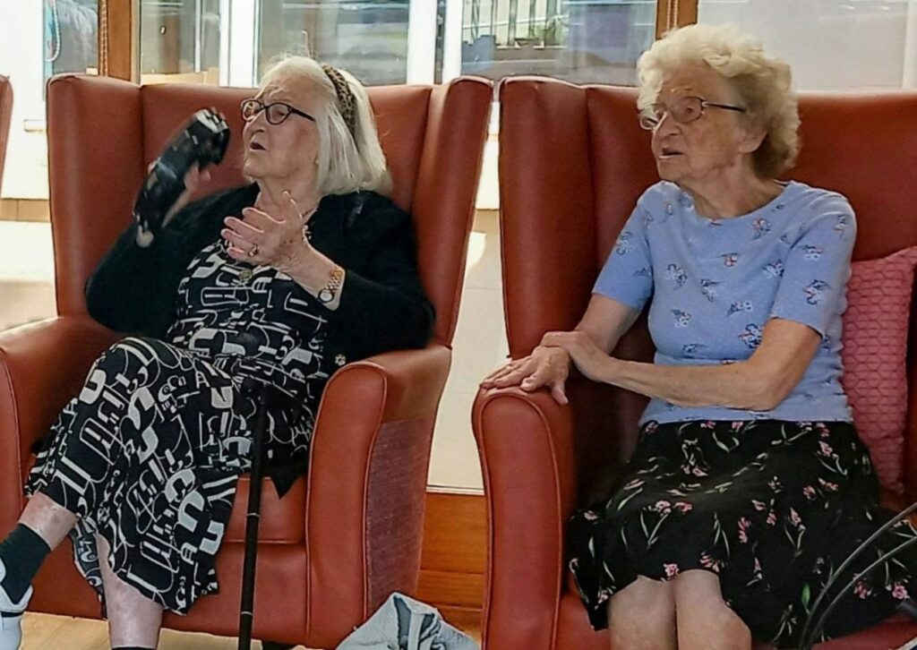 Two elderly women sit side by side in red armchairs. One gestures with her hand while holding a black object; the other has her hands clasped. Both seem engaged in conversation.