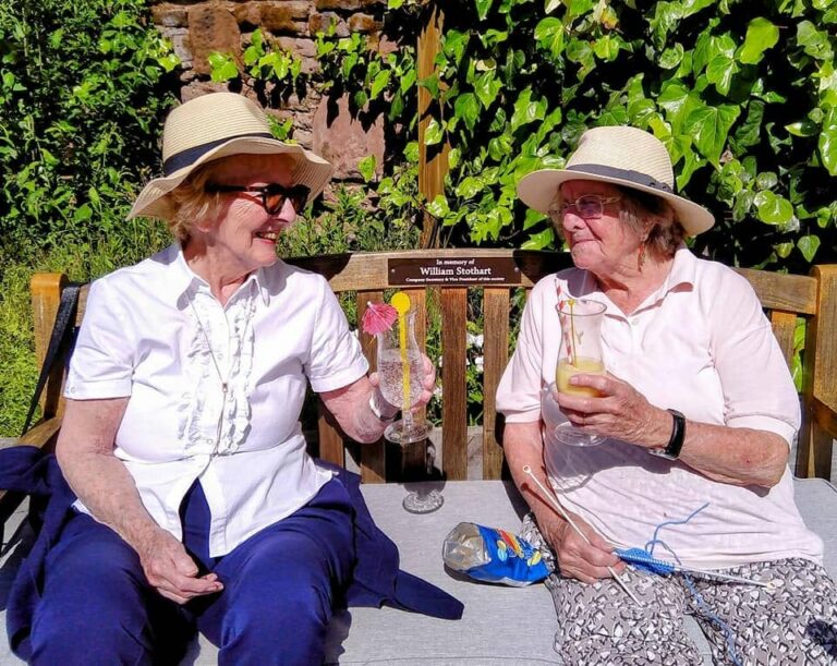 Two older women in hats sit on a bench outdoors, holding drinks and smiling at one another, enjoying the greenery and memorial plaque—a moment that captures the comfort of a welcoming home.