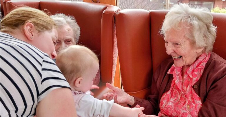 A smiling elderly woman reaches out to hold hands with a baby during intergenerational music time, whilst another elderly woman looks on in the background.