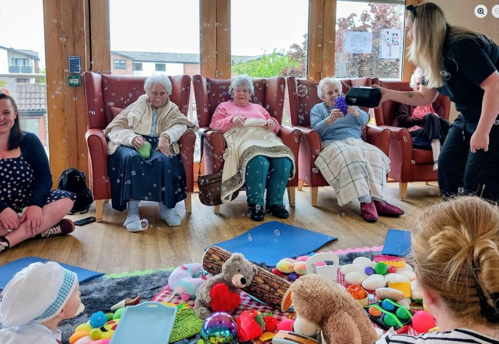 A group of elderly women sit on chairs watching a woman blow bubbles, with toys and play mats scattered on the floor before them during an intergenerational music and sensory play session.