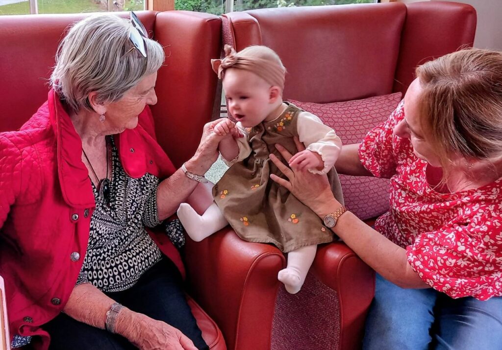 An elderly woman and an adult woman enjoy sensory play with a baby girl seated on a red armchair in a warmly lit room, sharing joyful Boogie Baby moments.