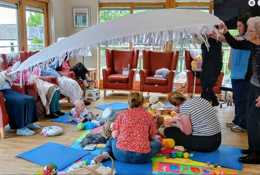 A group of adults and babies sit on mats in a brightly lit room, enjoying sensory play with toys as two people hold a large white fabric canopy overhead during a Boogie Baby session.