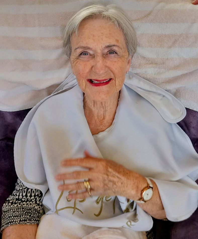 Elderly woman with short grey hair, wearing a patterned-sleeve top, a white serviette, a wristwatch and a gold ring, smiling at the camera whilst sitting on a striped towel.