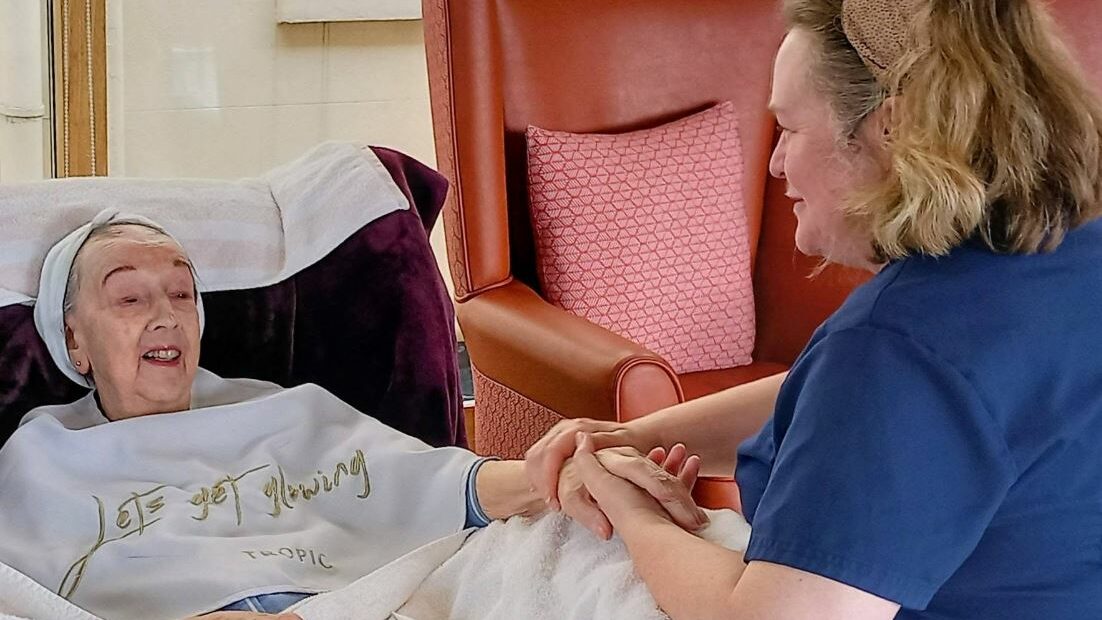 A carer in blue scrubs holds the hand of an elderly woman resting in a reclining chair, covered by a blanket that reads Lots of loving.