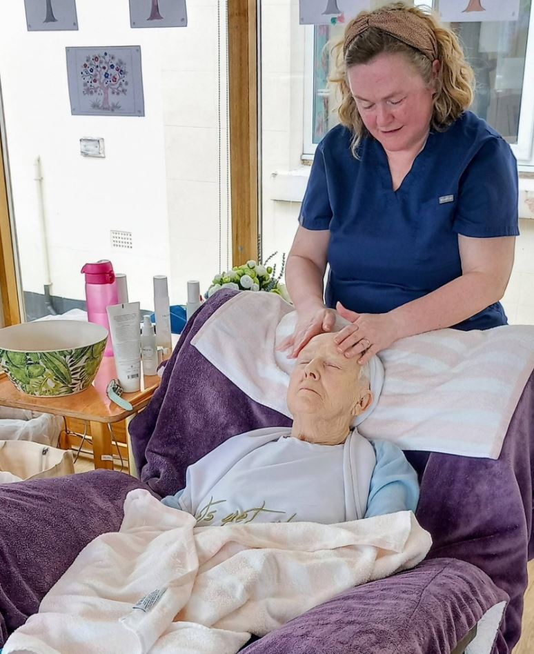 A woman in a navy uniform gives a facial massage to an elderly person reclining in a chair, surrounded by towels and skincare products.