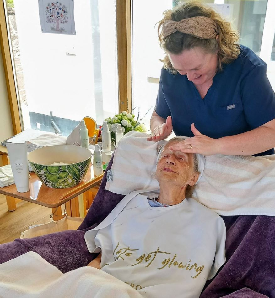A woman receives a facial treatment from a practitioner in a well-lit room, with skincare products and a bowl on a nearby trolley.